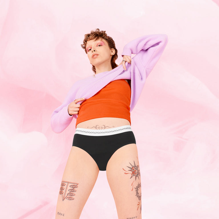 Femme en sous-vêtements tenant un pull, portant la culotte menstruelle ComfortFit Essentielle Hipster Logo Moderate, offrant protection et confort pendant les règles.